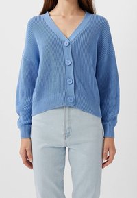 Blue knitted cardigan with a V-neck, ribbed texture, and five buttons. Features long sleeves and a cropped design, paired with light jeans.