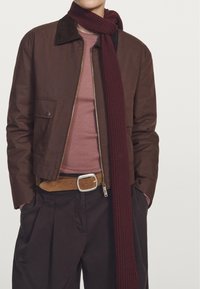 Brązowa kurtka z kołnierzykiem z korduropy, nałożona na różową bluzkę. W komplecie burgundowy szalik prążkowany, brązowy pasek i ciemne spodnie.