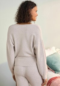 Svetlo siva pletena jopica z širokim izrezom in rebrastim robom, v kombinaciji s hlačami v enaki barvi. Ima spuščena ramena in mehak tekstil.