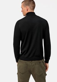 Schwarzer Strickpullover mit hohem Kragen, ausgestattet mit gerippten Bündchen und Saum. Kombiniert mit olivgrünen Hosen mit zwei Taschen hinten.