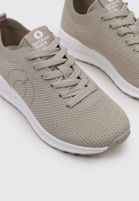 Zapatillas de punto beiges con un acabado texturizado, que presentan cordones planos, una suela acolchada y una etiqueta con el logotipo en el talón, con fondo blanco.