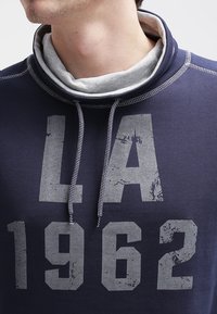 Felpa blu navy con dettagli grigi, collo avvolgente e grande stampa grafica grigia "LA 1962". Presenta un tessuto testurizzato e cordini.