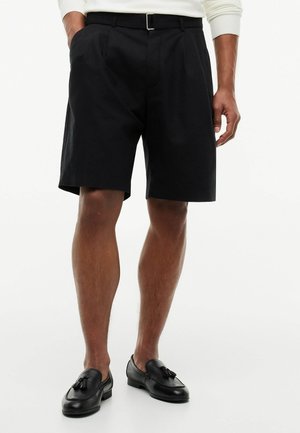 Mężczyzna w czarnych, szytych na miarę szortach z paskiem, białej bluzce z długim rękawem i czarnych skórzanych mokasynach z frędzlami, stojący na białym tle.