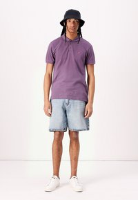 Camisa polo morada con cuello de botones, combinada con pantalones cortos de mezclilla claros y zapatillas deportivas blancas. Complementada con un sombrero tipo bucket negro.