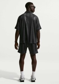 Άνδρας που στέκεται με την πλάτη, φορώντας μαύρο ριγέ πουκάμισο Nike, ταιριαστές σορτς, λευκές κάλτσες, λευκά αθλητικά παπούτσια και μαύρα γυαλιά ηλίου, μπροστά σε απλό φόντο.