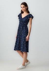 Navyblaues Kleid mit kurzen Rüschärmeln, floralem Muster, gerafftem Oberteil und knielangem Rock, kombiniert mit weißen Sneakers.