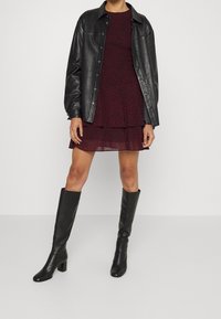 Giacca di pelle nera sopra un vestito bordeaux con un design a strati e stampa animale, abbinato a stivali neri con tacco al ginocchio.