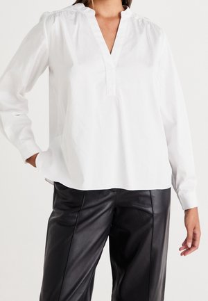 Femme portant un chemisier blanc à manches longues avec un col en V et un pantalon en cuir noir, debout avec une main dans la poche.