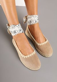 Chaussures espadrilles beiges avec bordure festonnée, tige en daim doux et semelle en jute tressée. Brides de cheville en tissu avec un motif floral noir.