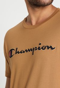 Hnědé bavlněné tričko s kulatým výstřihem, krátkými rukávy a vyšitým logem "Champion" v černé barvě s červeným akcentem. Hladká textura.
