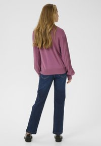 Mujer con cabello rubio largo, vistiendo un suéter de manga larga color malva y jeans oscuros, de pie de espaldas sobre un fondo blanco.