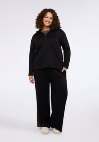 Sudadera con capucha negra con cremallera frontal y cordón, combinada con pantalones negros holgados con franjas laterales y zapatillas blancas.