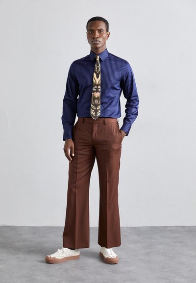 Chemise rayée marine avec une cravate à motifs, pantalon large marron, et baskets blanches avec des semelles beige. Tissu lisse et coupe ajustée.