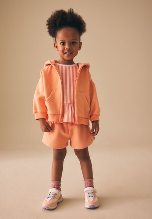 Jeune fille aux cheveux bouclés portant un sweat à capuche couleur pêche, une chemise rayée, un short assorti, des chaussettes roses et des baskets pastel, debout et souriante.