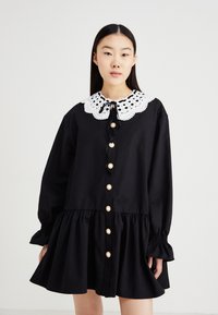 Rochie neagră din bumbac, cu guler din dantelă albă, mâneci lungi, închidere cu nasturi în față, cu embellismente florale și un tiv evazat.