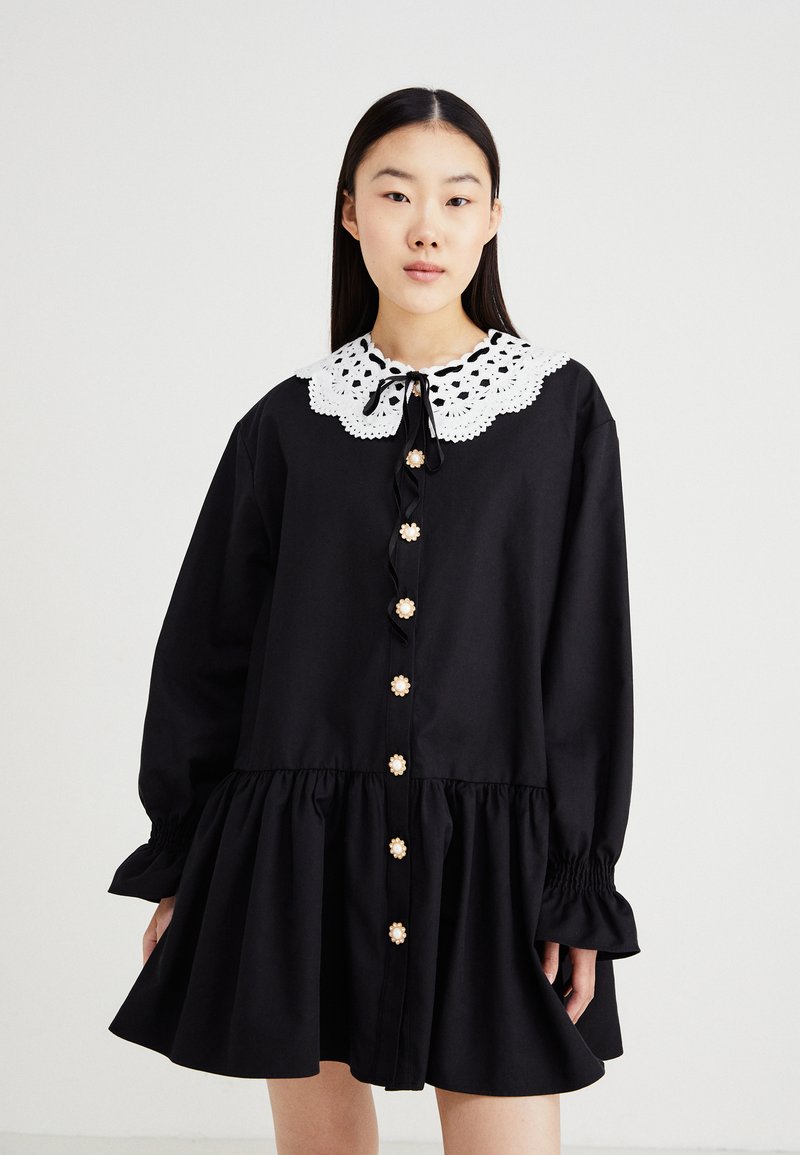 Rochie neagră din bumbac, cu guler din dantelă albă, mâneci lungi, închidere cu nasturi în față, cu embellismente florale și un tiv evazat.