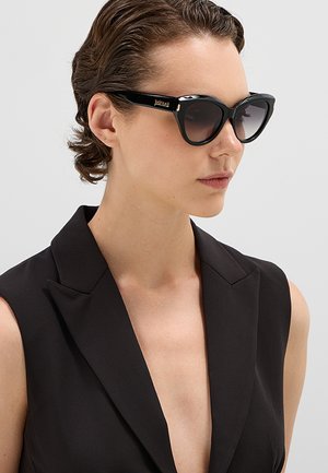 Donna con capelli corti e ricci indossa occhiali da sole neri a forma di occhio di gatto e un blazer nero senza maniche su pelle nuda su uno sfondo grigio chiaro.