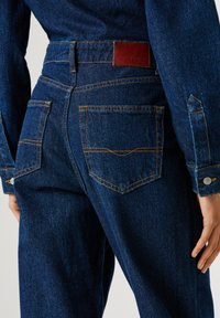 Džínsy z denimu s tmavomodrým umývaním, dizajnom s piatimi vreckami, kontrastným oranžovým prešívaním a hnedou koženou etiketou na zadnej strane.