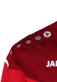Rote Sportjacke mit reflektierenden runden Punkten auf der Schulter und weißen Akzenten. Das Markenlogo ist gut sichtbar platziert.