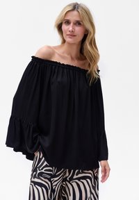 Schwarze schulterfreie Bluse mit elastischem Ausschnitt und gerafften Ärmeln, im lockeren Schnitt. Kombiniert mit gemusterten weiten Hosen.