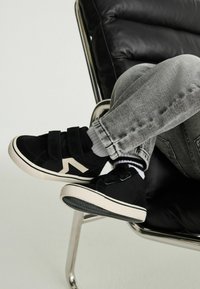 Svarta mockaskor med dubbel Veldorrem och en vit zigzagdetalj, ihop med grå jeans och randiga strumpor, på en svart stol.