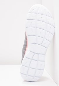 Semelle d'une chaussure de sport grise et rose avec motif de grip texturé blanc, positionnée verticalement contre une surface d'angle blanche.