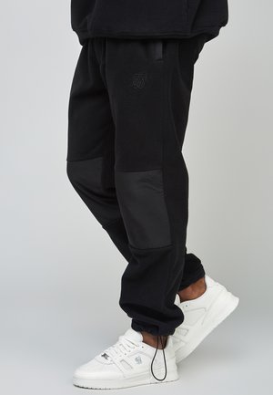 Person iført sorte fleece joggingbukser med forstærkede knælapper og hvide sneakers med logodetaljer.