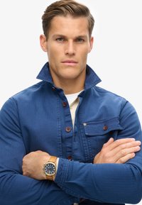 Camicia di jeans blu con colletto alto, chiusura con bottoni e tasca dettagliata. Al polso è indossato un orologio da polso in pelle marrone con quadrante blu.