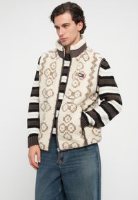 Fleecová vesta v krémovej farbe s hnedým kvetinovým vzorom, so zipsom, vysokým golierom a bočnými vreckami. Nosená cez pruhovaný hnedo-krémový sveter.