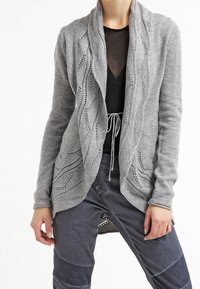 Grå stickad kofta med draperad halsringning och öppen front. Har ett texturerat mönster längs kanterna och ribbade manschetter.