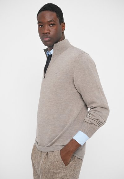 Beige félzipperes pulóver bordás mandzsettákkal és alsó résszel, világoskék ing fölött viselve. Texturált anyaggal és diszkrét logó részlettel rendelkezik.