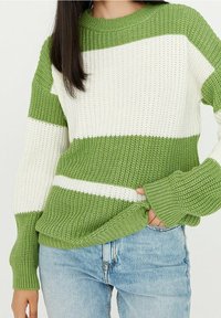 Donna che indossa un maglione di maglia a righe verdi e bianche con maniche svasate, infilato leggermente nei jeans azzurri, con una mano nella tasca.