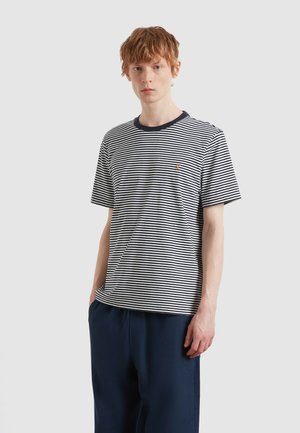 Młody mężczyzna o rudych włosach, ubrany w granatowo-białą pasiastą koszulkę z krótkim rękawem i ciemnoniebieskie spodnie, stoi na tle jednolitego tła.