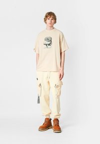 Joven vestido con una camiseta gráfica de gran tamaño beige, pantalones cargo color crema con bolsillos con cremallera y botas marrones con cordones, de pie sobre un fondo blanco.