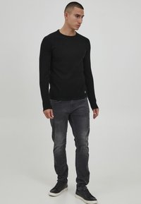 Zwarte langemouwen sweater met een gestructureerd design, gecombineerd met grijze jeans en zwarte sneakers met een witte zool. Schoon en minimalistisch stijl.