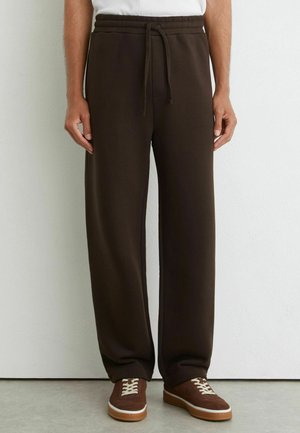Mand iført mørkebrune sweatpants med snøre og brune sneakers, der står op ad en hvid væg.