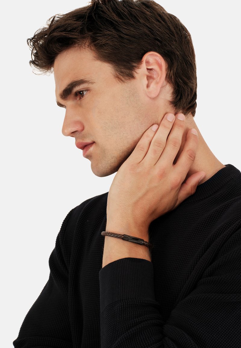 Joven con suéter negro tocándose el lateral del cuello, mostrando una pulsera de cuero trenzado con cierre de metal en la muñeca, fondo claro.