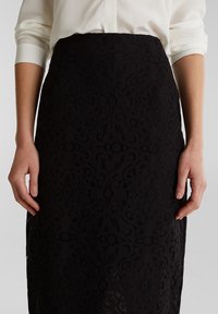 Gonna nera in pizzo con intricate fantasie floreali. Un design aderente con una superficie liscia, che presenta una vita ben definita e un taglio a lunghezza ginocchio.