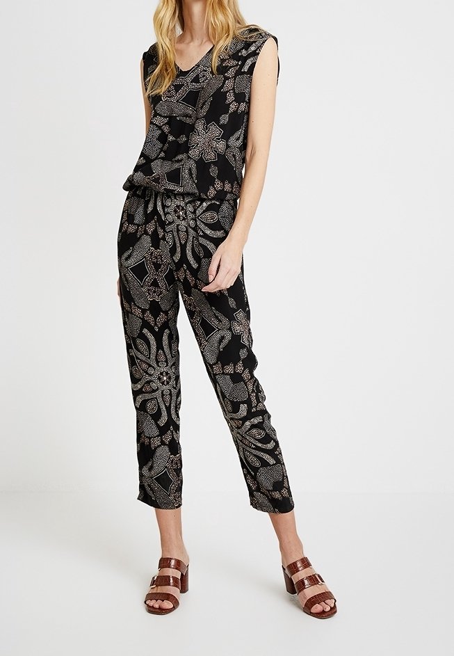 Schwarzes Outfit mit komplexen silbernen und goldenen Blumendrucken, ärmelloses Top mit geschichtetem Design und taillierten Hosen; getragen mit braunen Sandalen.