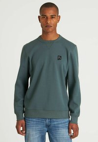 Sudadera verde con mangas largas, cuello redondo, tela texturada y un pequeño parche negro con el logo en el lado izquierdo del pecho. Llevado con jeans azules.