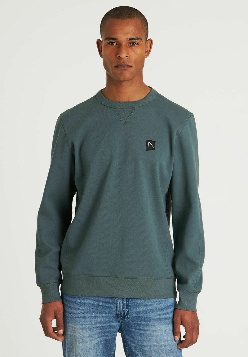 Sudadera verde con mangas largas, cuello redondo, tela texturada y un pequeño parche negro con el logo en el lado izquierdo del pecho. Llevado con jeans azules.