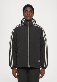 Črna podložena jakna z visokim ovratnikom, belimi stranskimi črtami, sprednjo zadrgo in elastiko na manšetah. Na spodnji levi strani ima logotip v obliki značke.