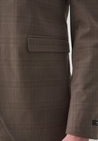 Blazer cu carouri maro, realizat dintr-un material texturat, având un buzunar exterior, umeri structurați și un design ascuțit al reverului.