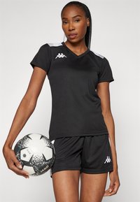 Čierny športový dres s krátkymi rukávmi a V-výstrihom, s bielymi akcentmi a logom. V kombinácii s čiernymi šortkami a drží čiernobiely futbalový loptu.