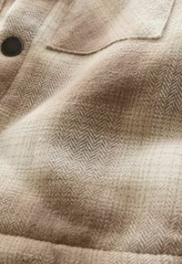 Tkanina w wzór houndstooth w odcieniach beżu i kremu, charakteryzująca się miękką teksturą z wykończeniem szczotkowanym i detalem w postaci guzika.