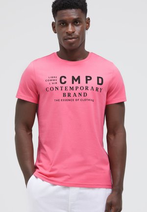 Junger Mann trägt ein rosa T-Shirt mit schwarzem Text und weißen Hosen, steht vor einem schlichten hellgrauen Hintergrund.