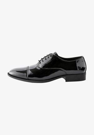 Chaussure Oxford en cuir noir brillant avec bout droit, laçage fermé et petit talon, vue de profil sur fond blanc.