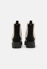 Black suede ankle boots with white faux fur lining, thick black rubber soles, and back pull tabs. Features a gold logo detail at the heel.