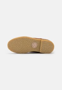 Brown suede shoe sole with textured rubber grip. Features a circular brand logo on a light pink disc near the heel.