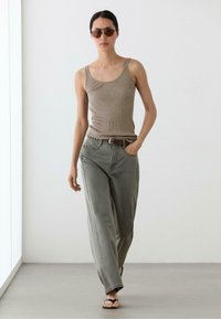 Femme portant un débardeur côtelé marron, pantalon large gris avec ceinture, tongs noires et lunettes de soleil marron marchant dans un espace intérieur minimaliste.
